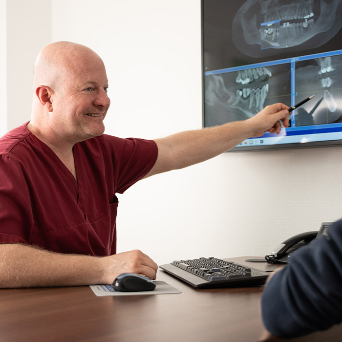 Dr. Roers sitzt mit Patienten am Tisch und zeigt auf Röntgenaufnahme auf Bildschirm an der Wand.